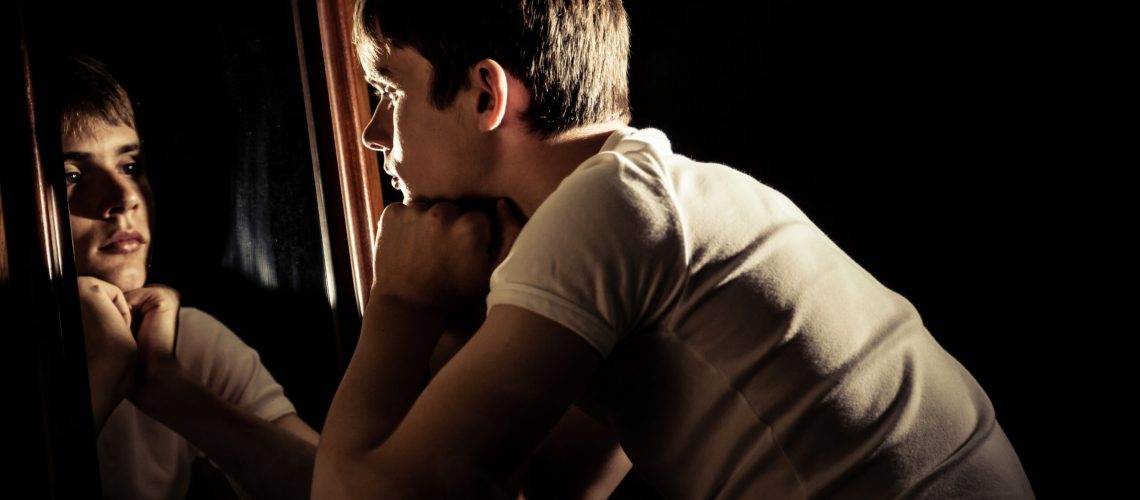 Serious teen boy surrounded by darkness looking at himself in mirror with wooden frame